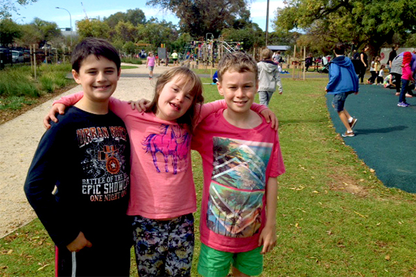 3 primary school aged children at a park, smiling, with their arms draped around each other's shoulders
