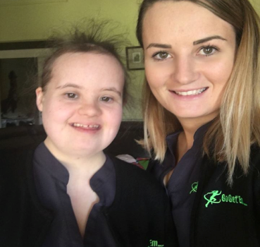 Two young women smiling at the camera, wearing black shirts with a green logo