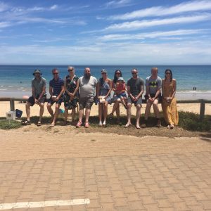 a group of 9 adults and one young child sit casually along a fence in front of a beach on a sunny day.