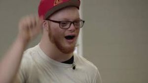 A man with a red beard, white t-shirt red flat brimmed cap and glases is speaking, with his fist waving in the air in an affirming position
