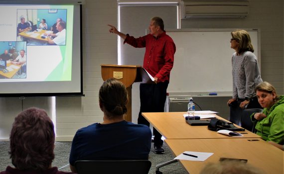 A tall man wearing dark pants and a red shirt stands at the front of a room talking to people seated and looking at a presentation on a screen