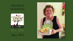 A green background presentation screen with logo of an illustrated tree with coloured leaves. Photo on the right of a woman in a black sleveless top holding a box of packaged dried fruit. She has chin length curly dark blonde hair and is smiling at the camera.