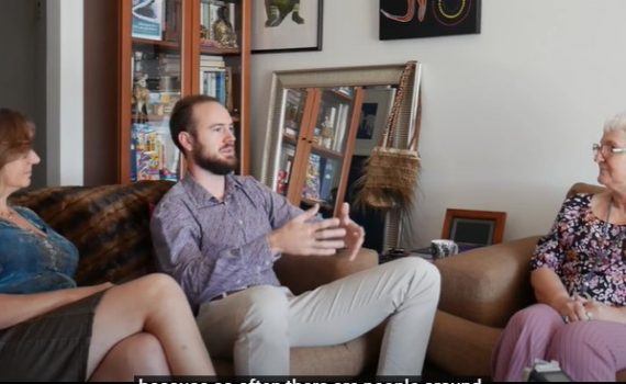 Two adult women and a male in conversations, sitting on chairs in a lounge room with a bookcase behind them