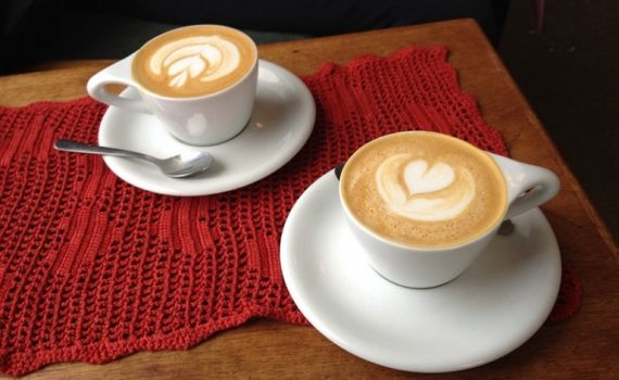 Two cups of coffee with heart shapes made in the top of the milk froth. The cups are on white saucers and on a red cloth on a wooden table