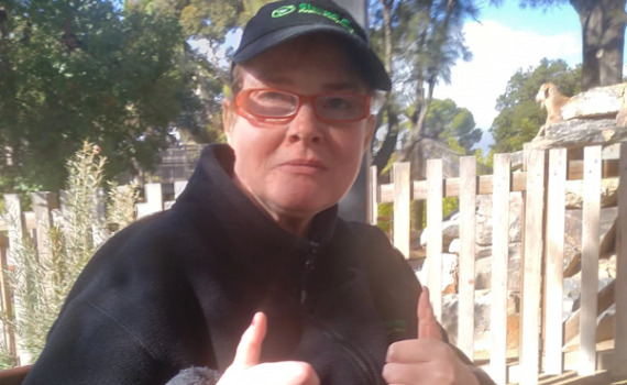 A woman gives 2 thumbs up to the camera. She is outdoors, wearing a black cap and jumper with a green shred em logo and has glasses with red frames
