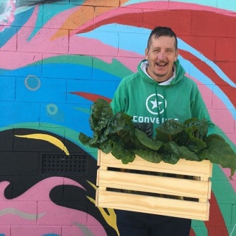 A tall man in a green hoodie is smiling and holding a large wooden crate filled with green leafy vegetables. He is standing in front of a brightly painted wall mural