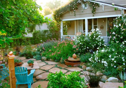 Grey and white painted porch of house visible behind a lush green garden with whte flowers and brown pavers.