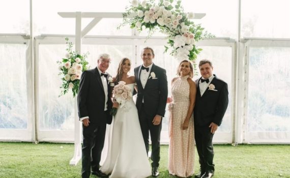 Wedding photo father, bride, groom, mother, brother in elegant formal clothing standing on grass in front of a white wooden frame adorned with a large bunch of roses