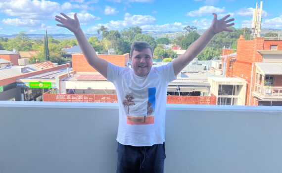 A young adult, standing on an apartment balcony, arms outstretched, suburban rooftops and blue sky in the background.