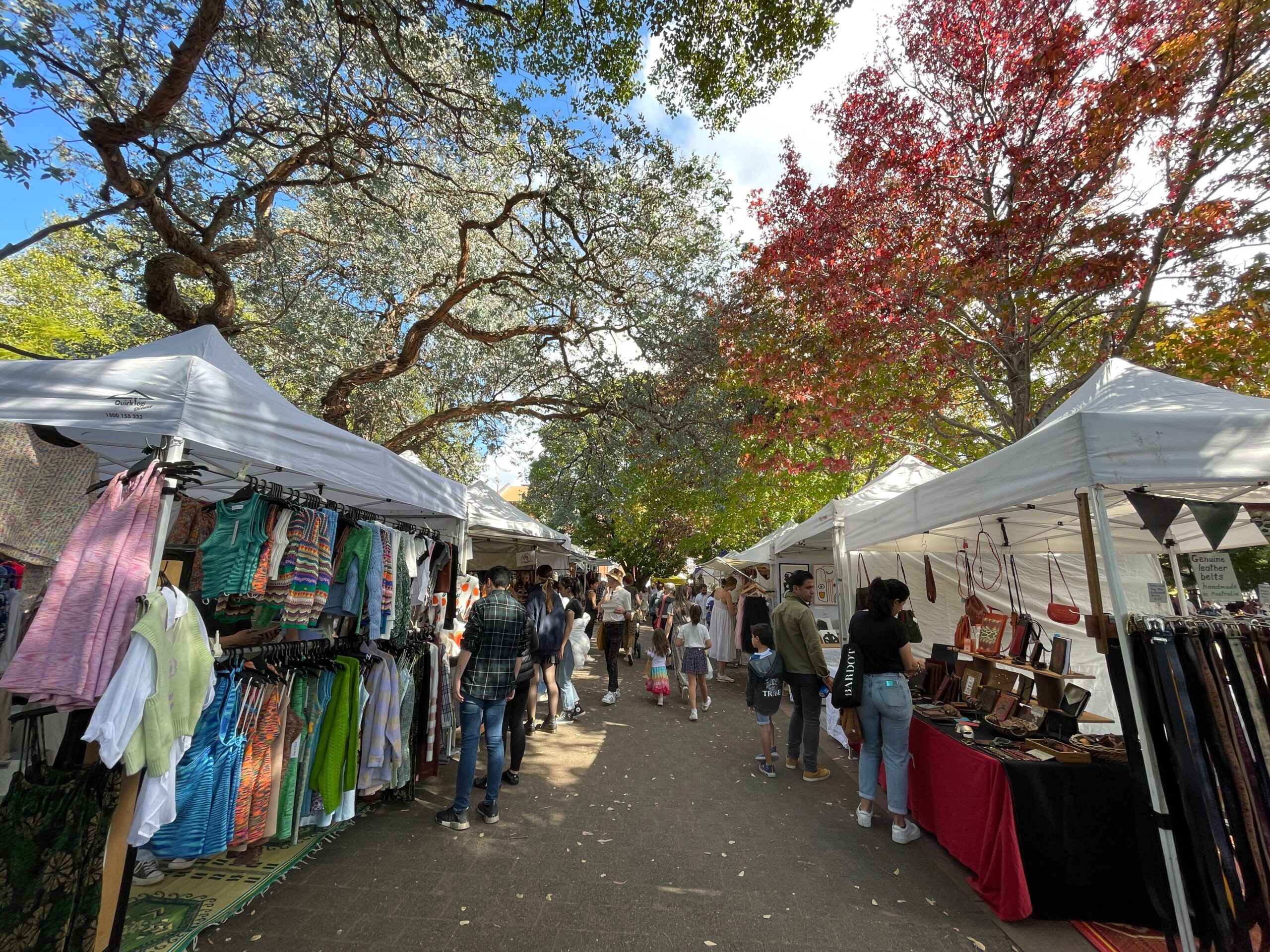 An outdoor market - a walkway between two rows of white tents with crafts and clothing on display. overhead there are large trees with purple, green and autumn coloured leaves