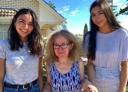 Three women standing together shoulder to shoulder, with warm smiles. In the centre is a woman with curly blonde hair and glasses. By her side are two younger women with a family resemblance in their dark hair and olive skin. They are standing in front of a neat brick home on a sunny day.