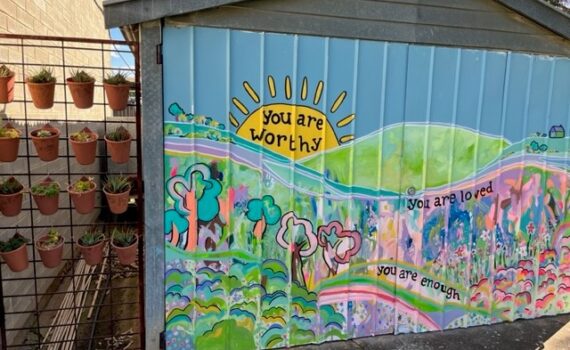 A mural painted on a shed wall. There is a village scene, mountain behind and a bright yellow sun with the words You are worthy