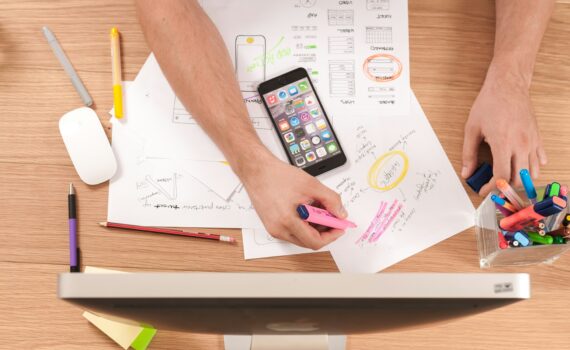 looking down on a table - a person is brainstorming on a piece of paper with coloured pencils, there are circled and highlighted notes on a page, their phone is laid on top and there is a computer monitor in view.