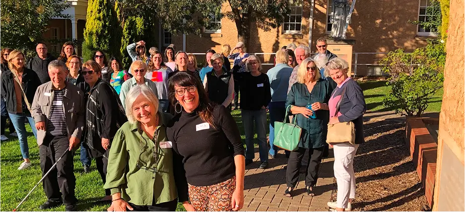 A group of 30 people are gathered on a lawn in the early morning sun. They are gathered in a loose group but have been caught off-guard, many leaning in together talking in groups of two. Most are looking up and smiling, some holding their hand up to shield their eyes from the sun