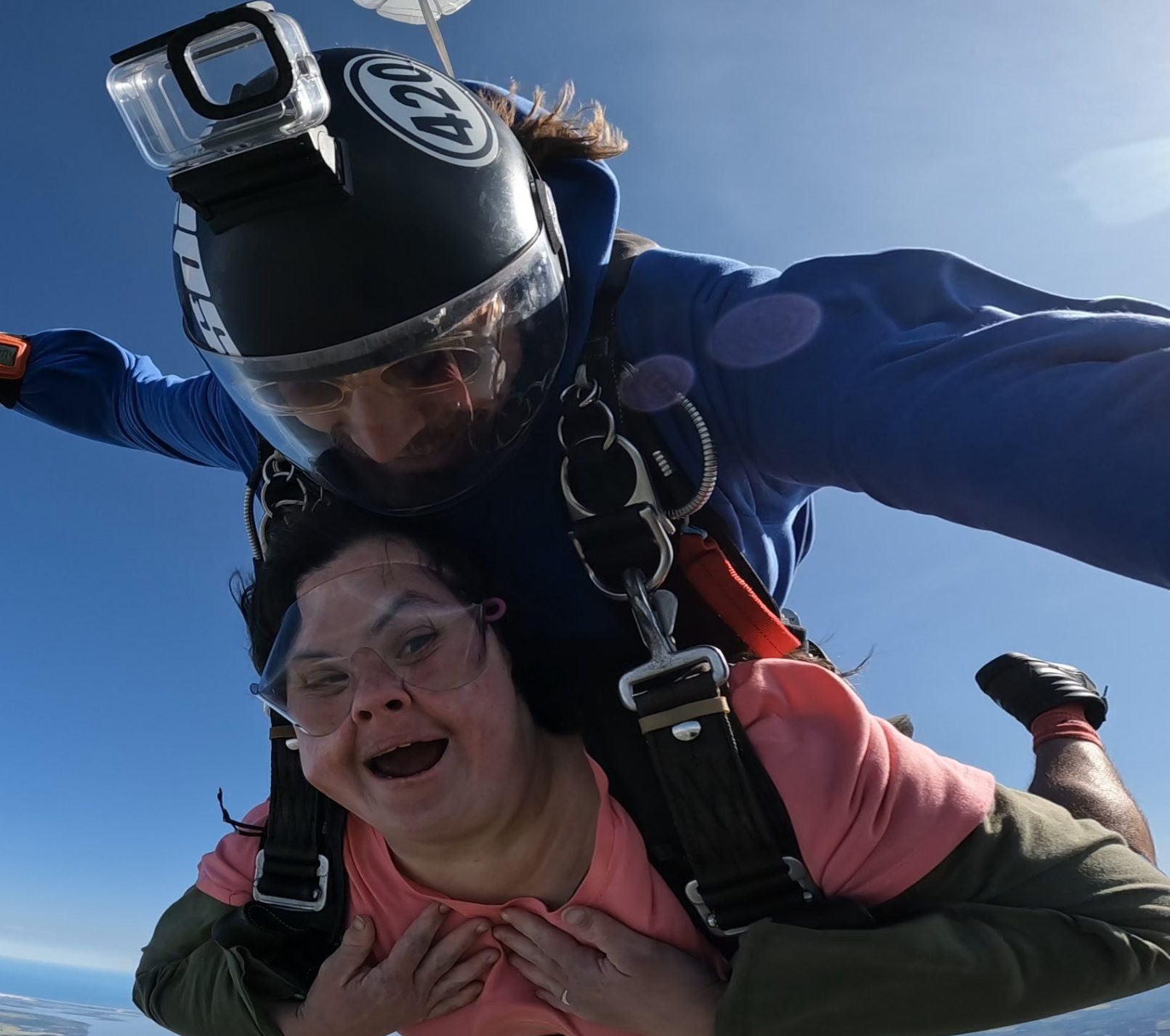 A woman is in a tandem skydive - strapped to an instructor above her with his arms stretched wide. She has her mouth open smiling and the wind has caught her cheeks. Her hands are touching her chest