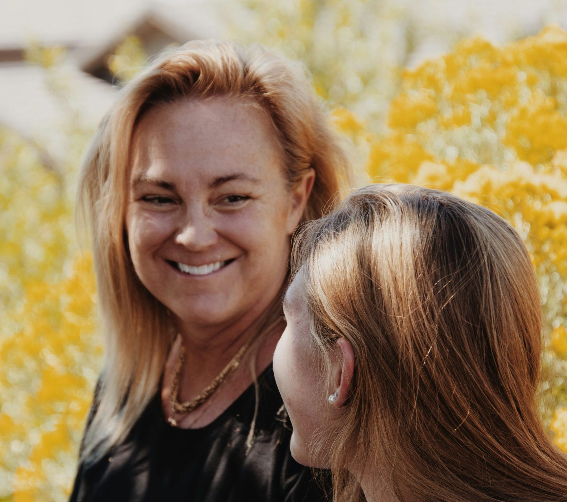 Two middle aged women with strawberry blonde hair look at each other smiling. One is facing the camera, the other has her back to the camera. Bushes in the background are in autumn colours.