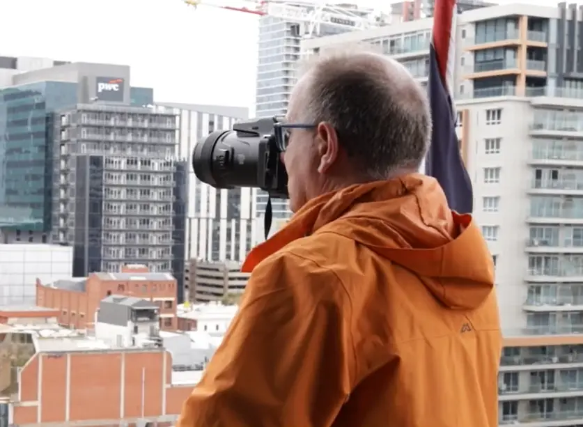 A photographer is looking through a camera with a long lens. He's up high looking across city buildings.