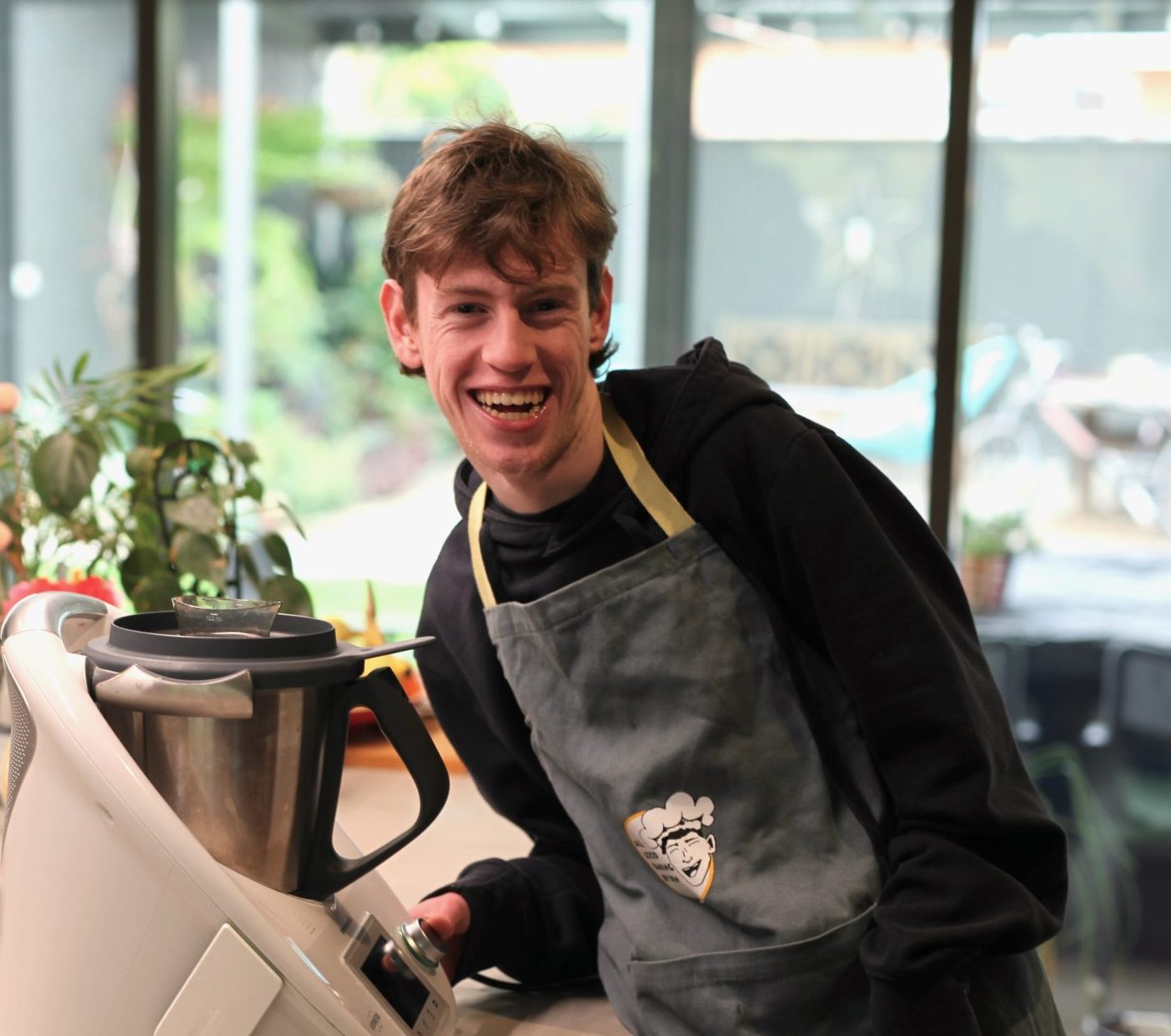 A young adult man is at a kitchen bench next to a mixer. He's smiling at the camera and wearing a professional apron with a cartoon in his likeness printed on the front