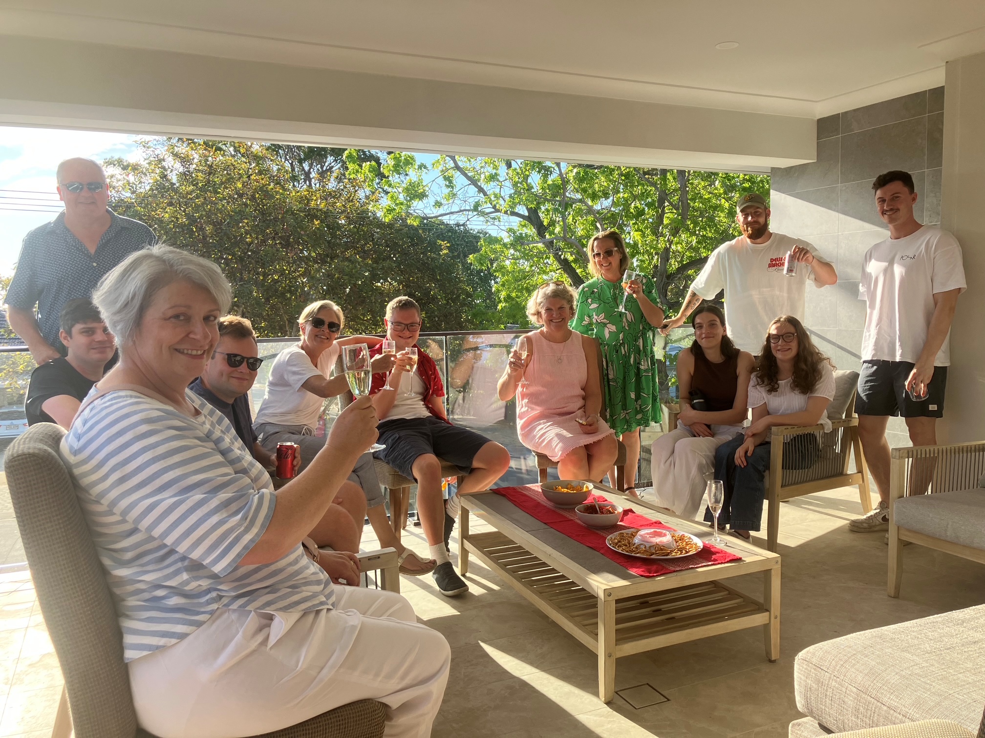 A group of 12 adults in a festive setting on a balcony in the Australian Sun. They are holding up glasses of champagne to say 'cheers'. There are plates of cheese and nibbles in platters on a red table runner.