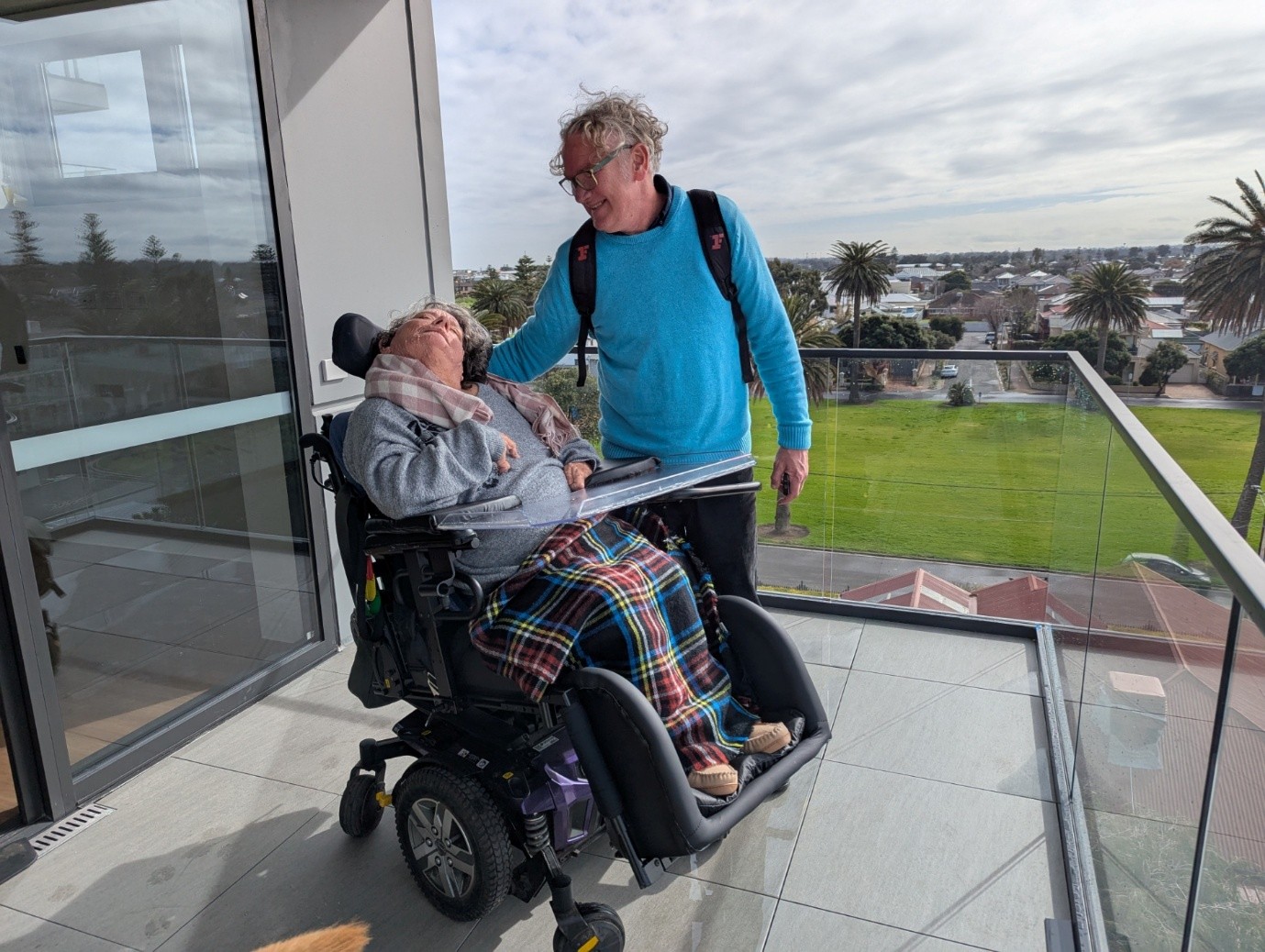 An adult woman and man are in conversation on an apartment balcony. There are lawns and palm trees in the background. She's seated in a powered wheelchair and has a got a tartan rug over her knees and a scarf around her neck. The man she's speaking with is wearing a bright blue jumper and a backpack.