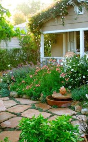 Grey and white painted porch of house visible behind a lush green garden with whte flowers and brown pavers.